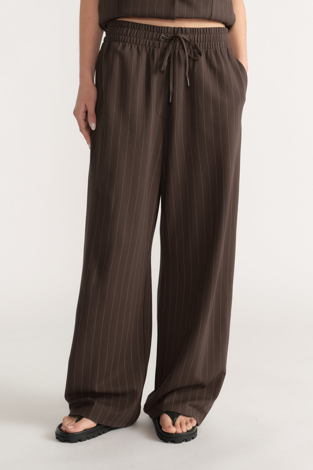Wide-leg dark brown pants with white pinstripes, elasticated waist, drawstring, and side pockets worn with sandals