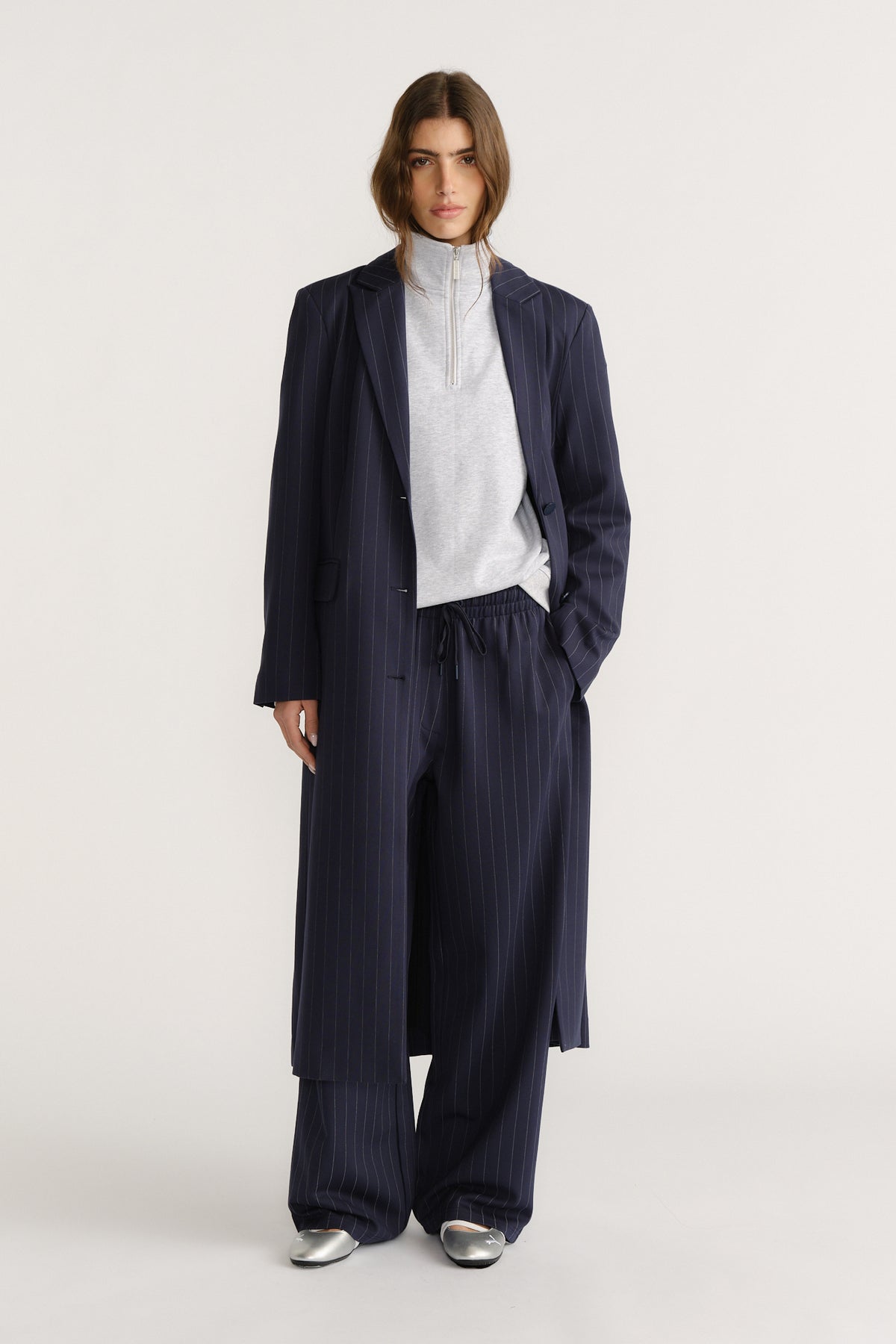 Woman wearing navy pinstripe wide-leg pants and matching long blazer layered over a light grey zipped pullover