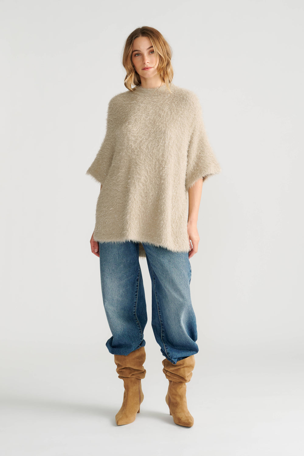 Woman wearing a relaxed oversized knit top in soft biscuit tone paired with loose blue jeans and slouchy tan boots.