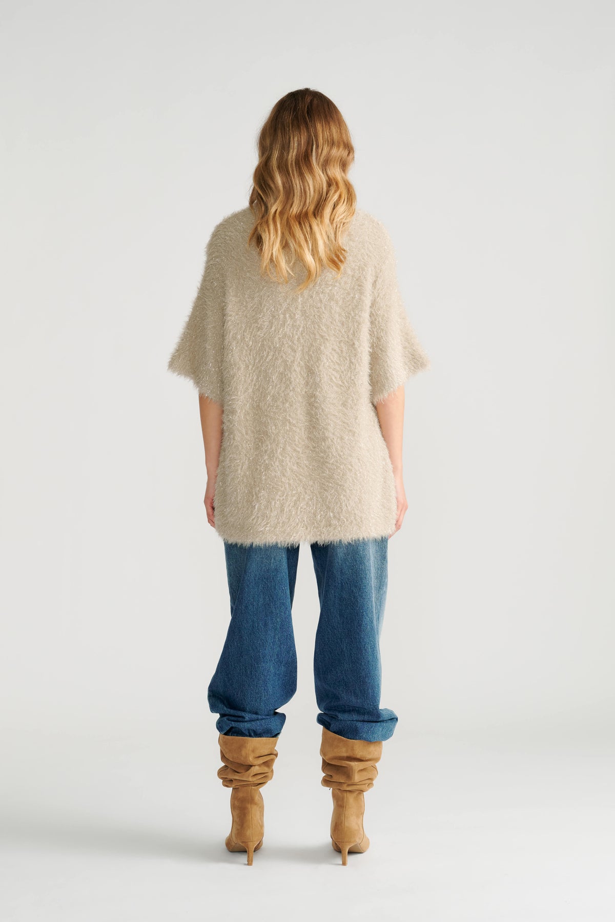 Woman wearing a relaxed biscuit-colored oversized knit tee paired with loose blue jeans and slouchy tan boots, back view.