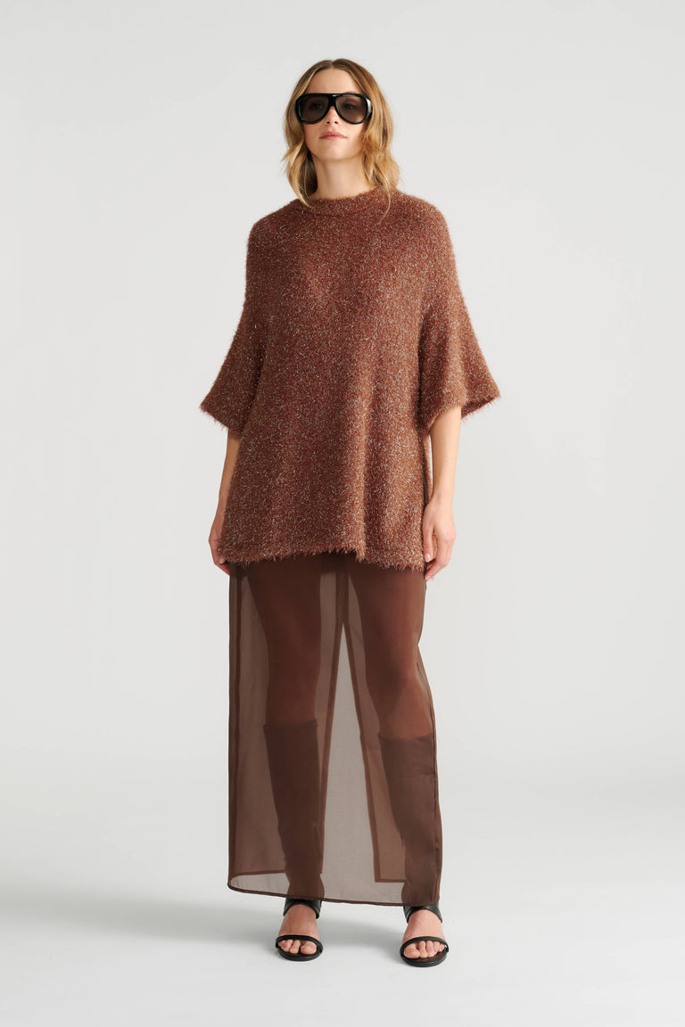 Woman wearing a textured oversized knit top in warm brown tones paired with sheer wide-leg pants and black heels.