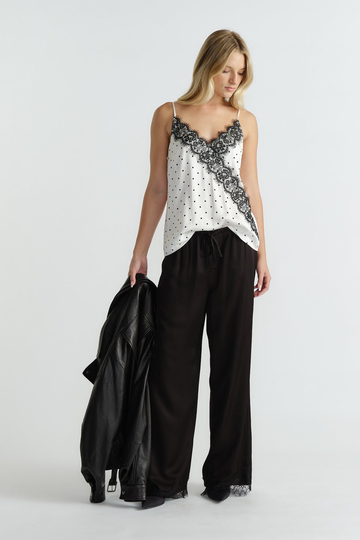 Woman wearing ivory lace cami with black polka dots paired with relaxed black pants, holding a black leather jacket.