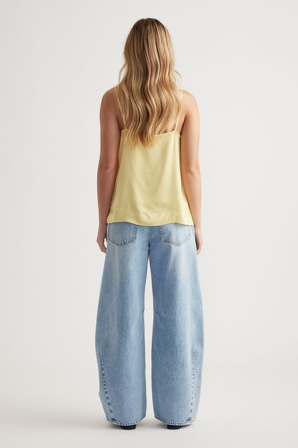 Woman with loose wavy hair wearing a soft yellow lace cami paired with light blue wide-leg denim jeans viewed from behind