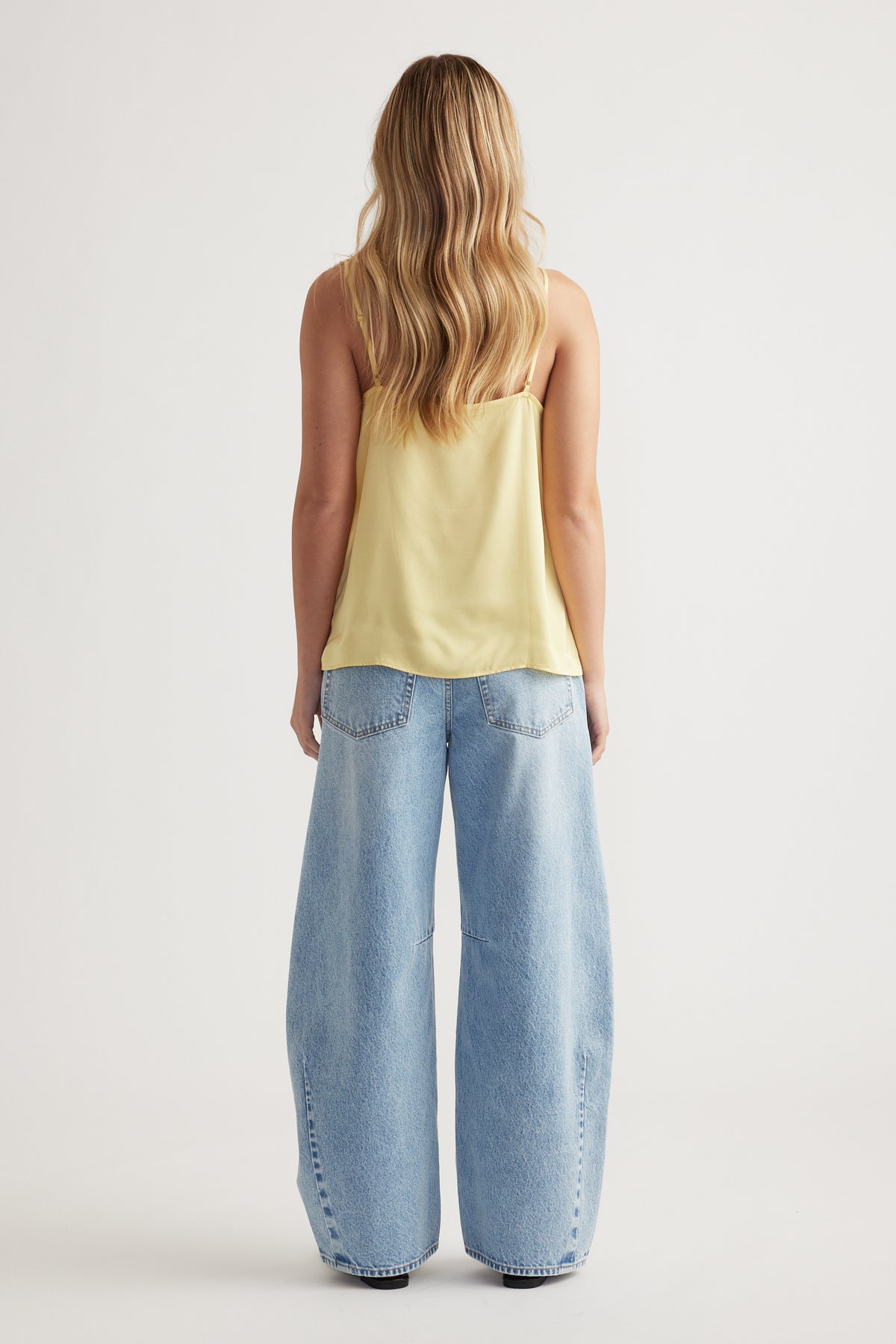 Woman with loose wavy hair wearing a soft yellow lace cami paired with light blue wide-leg denim jeans viewed from behind