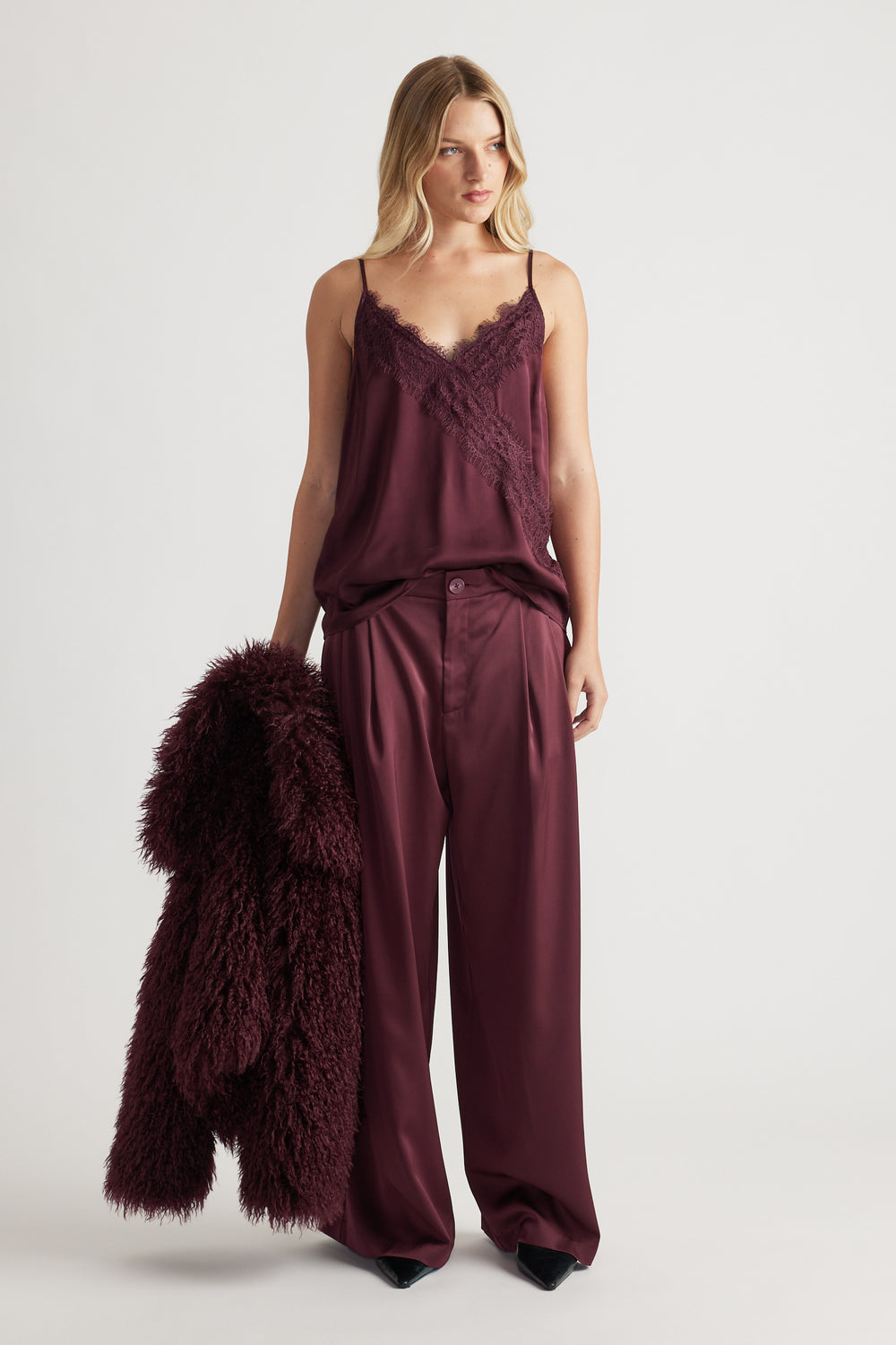 Woman wearing a plum lace-trimmed cami paired with wide-leg satin trousers, holding a matching textured jacket for a luxe look