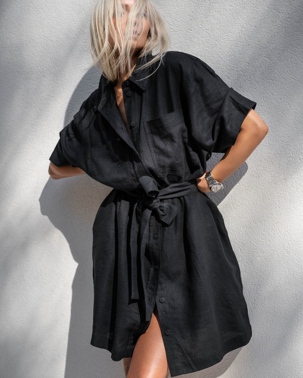 Woman wearing a black linen blend short dress with a belted waist and rolled sleeves standing against a textured wall.