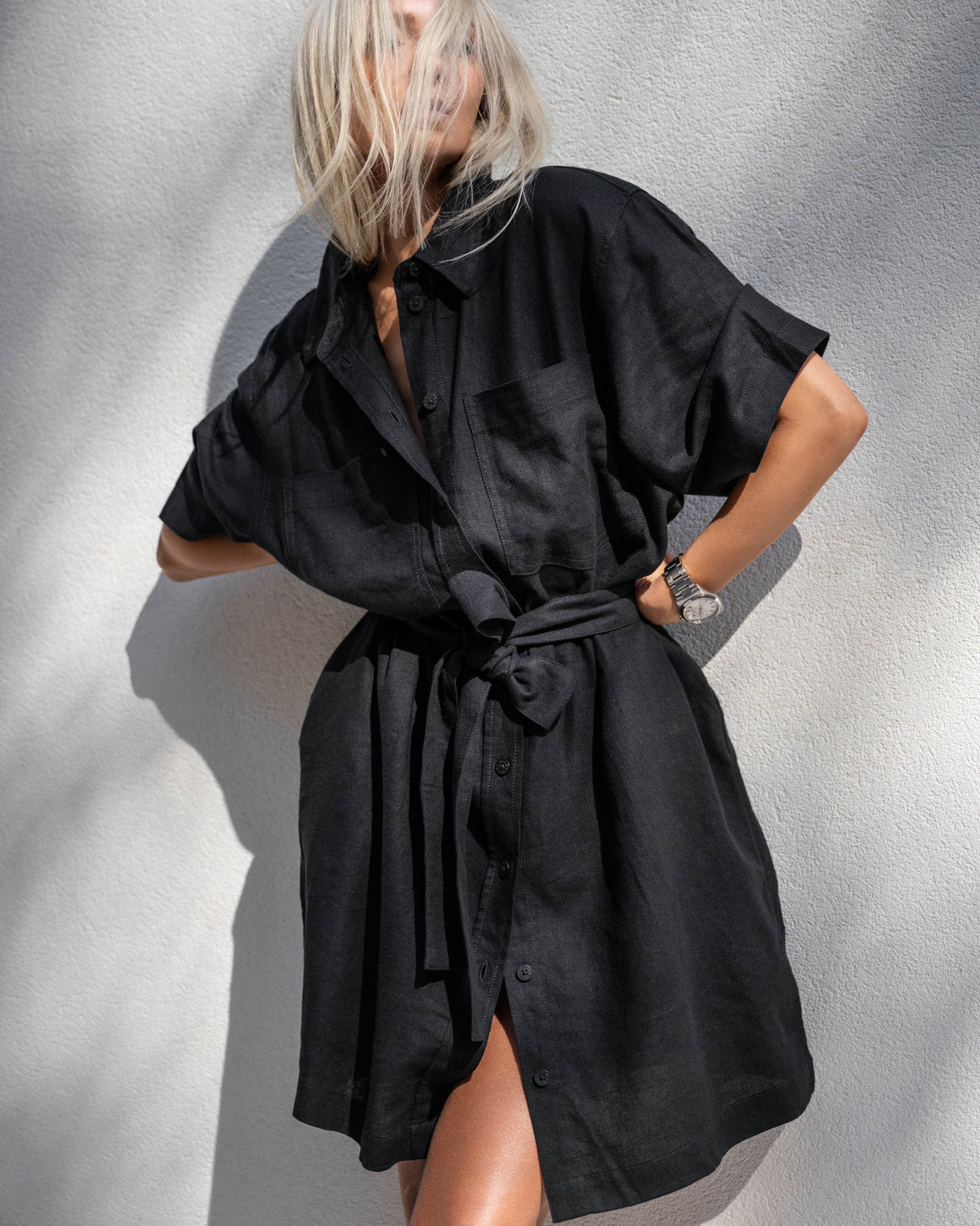 Woman wearing a black linen blend short dress with a belted waist and rolled sleeves standing against a textured wall.