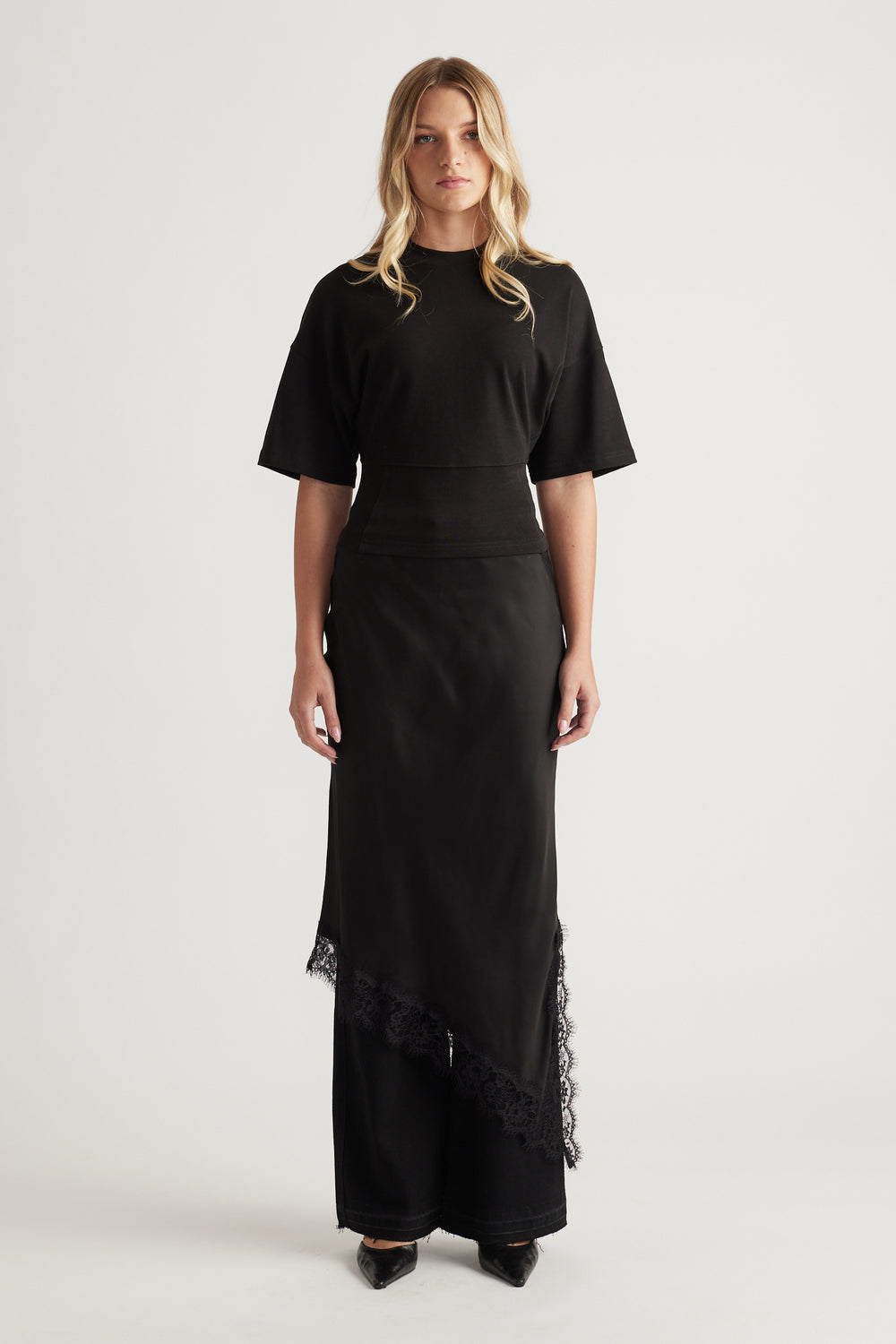 Woman wearing a black fitted waist tee paired with a long asymmetrical skirt featuring lace trim and pointed black shoes
