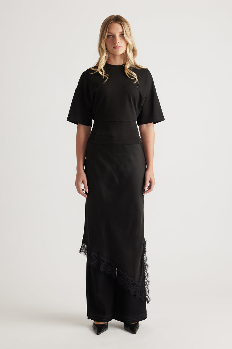 Woman wearing a black fitted waist tee paired with a long asymmetrical skirt featuring lace trim and pointed black shoes