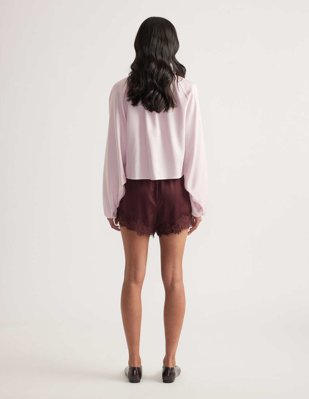 Back view of a woman wearing a light pink collared jacket with puffed sleeves, burgundy shorts and black loafers