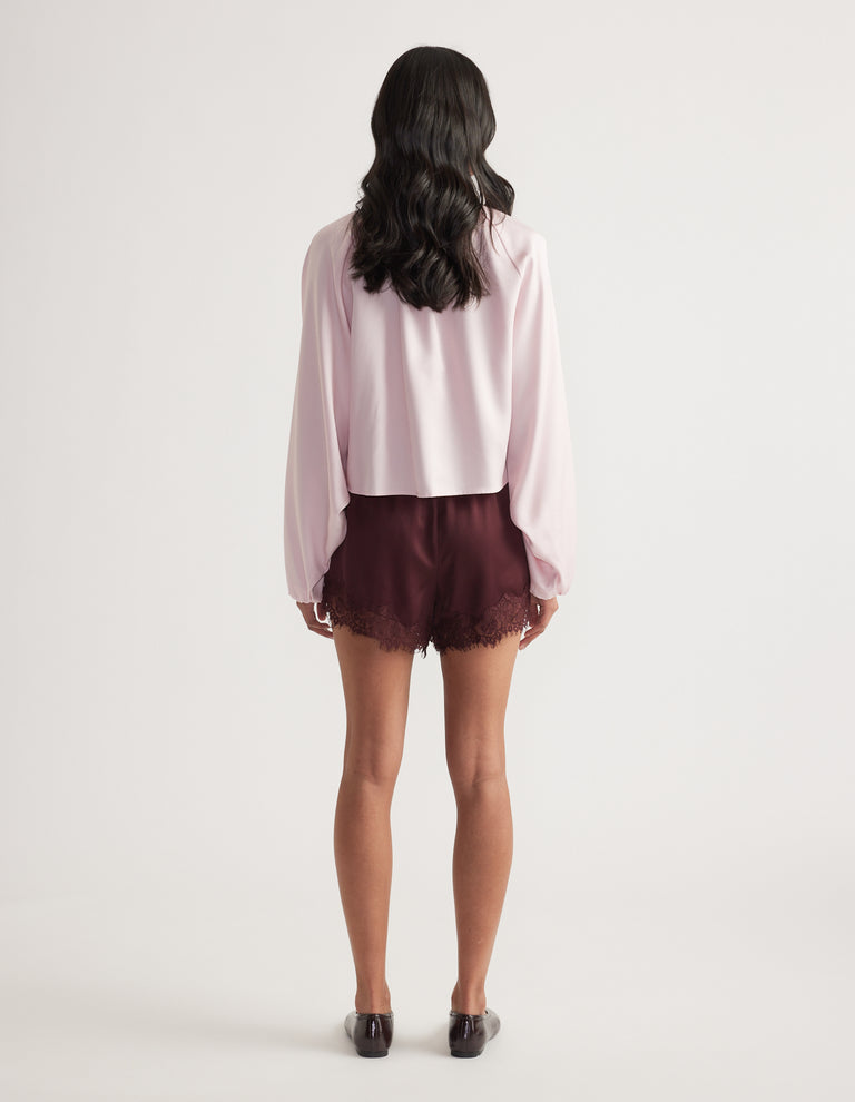 Back view of a woman wearing a light pink collared jacket with puffed sleeves, burgundy shorts and black loafers