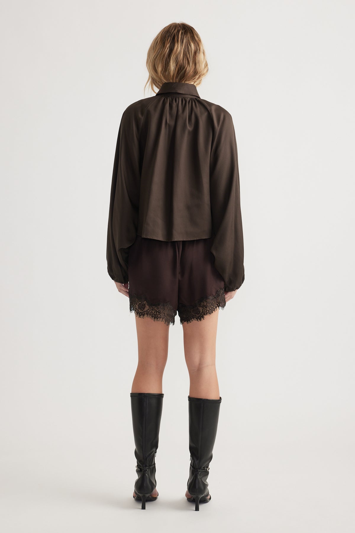 Woman wearing a loose dark long-sleeve top paired with black lace-trimmed shorts and knee-high boots, back view