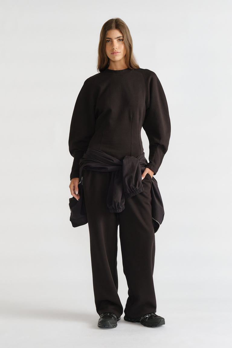 Woman wearing a fitted black cinched sweatshirt with puff sleeves and loose black pants styled with a tied jacket at the waist
