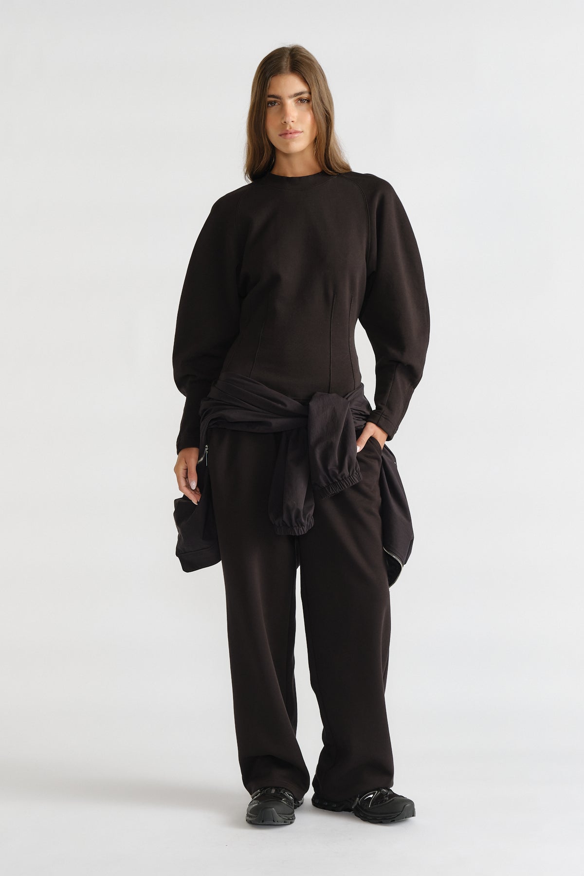 Woman wearing a black cinched waist sweatshirt with balloon sleeves, paired with loose black pants and sneakers.