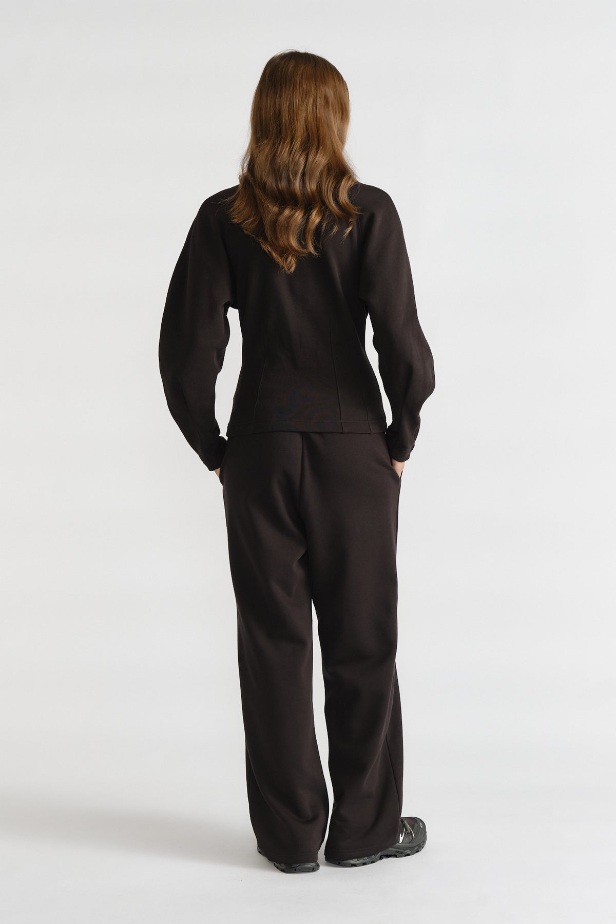 Woman wearing relaxed black cinched sweatpants and matching long-sleeve top with hands in pockets, standing facing away