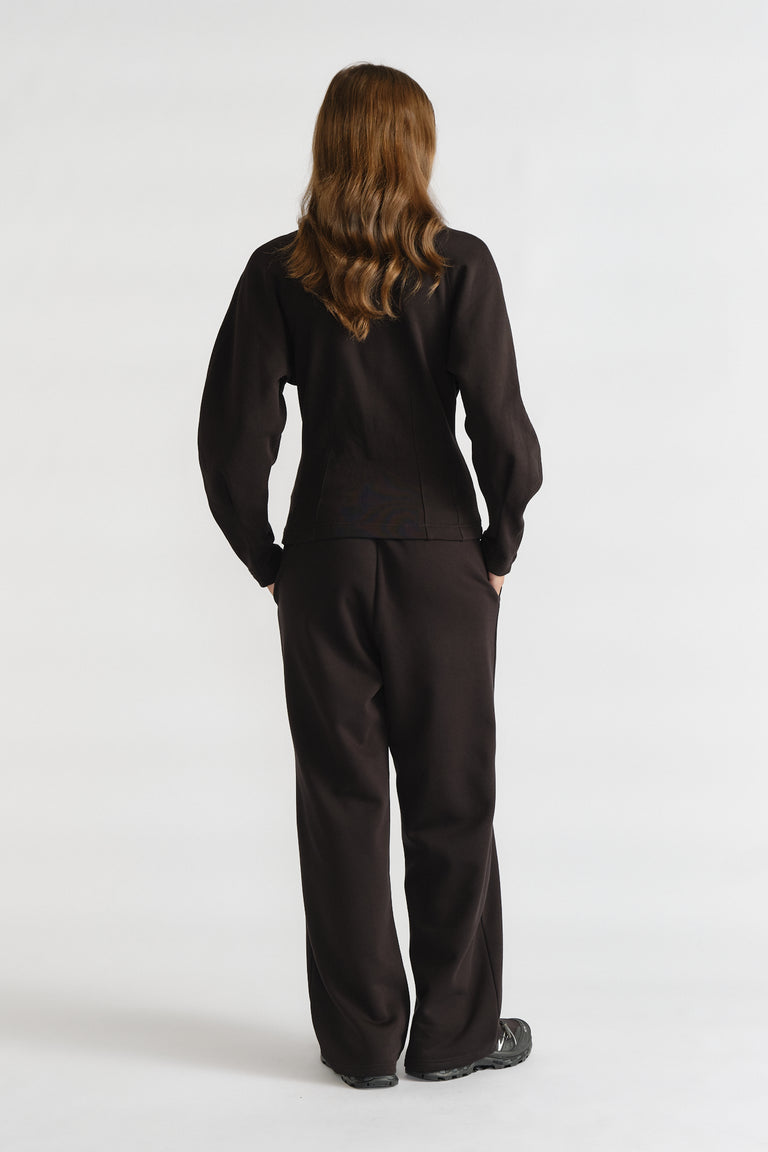 Woman wearing relaxed black cinched sweatpants and matching long-sleeve top with hands in pockets, standing facing away