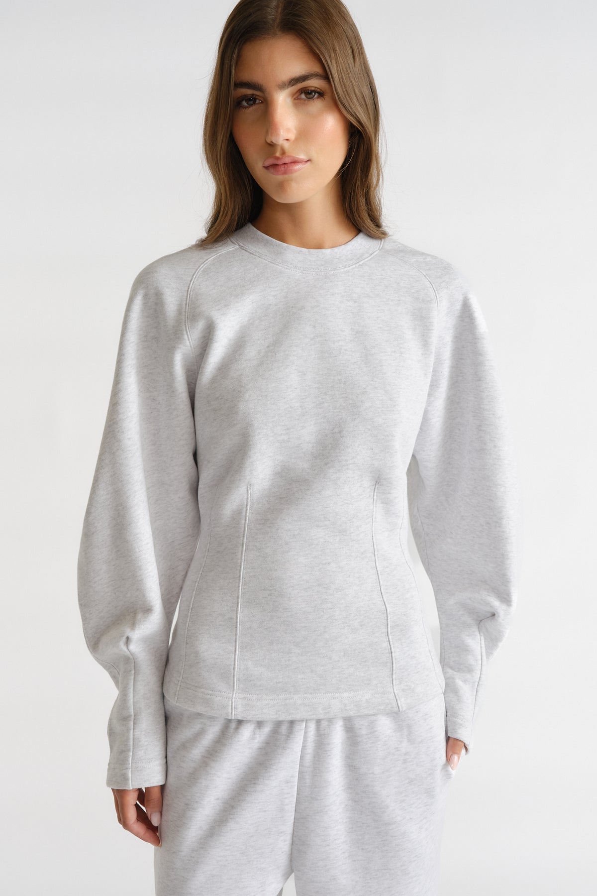 Woman wearing light grey cinched sweatshirt with long balloon sleeves and matching sweatpants against plain background