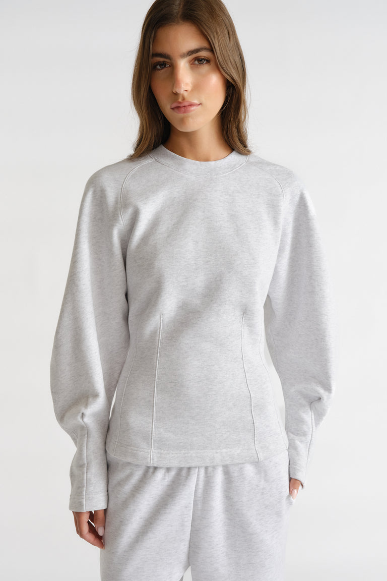 Woman wearing light grey cinched sweatshirt with long balloon sleeves and matching sweatpants against plain background