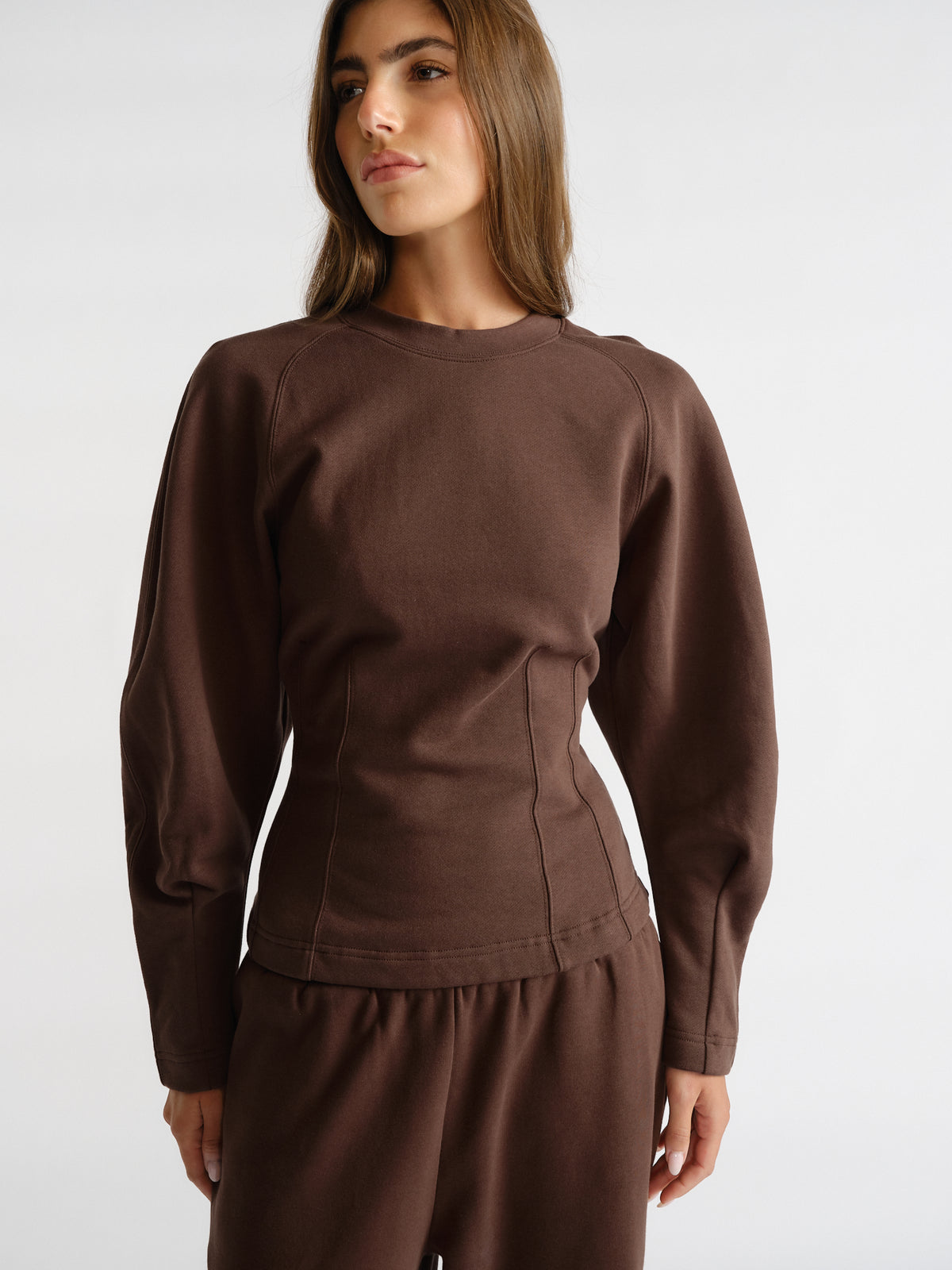 Woman wearing a rich brown cinched waist sweatshirt with long sleeves, paired with matching relaxed pants on a white background
