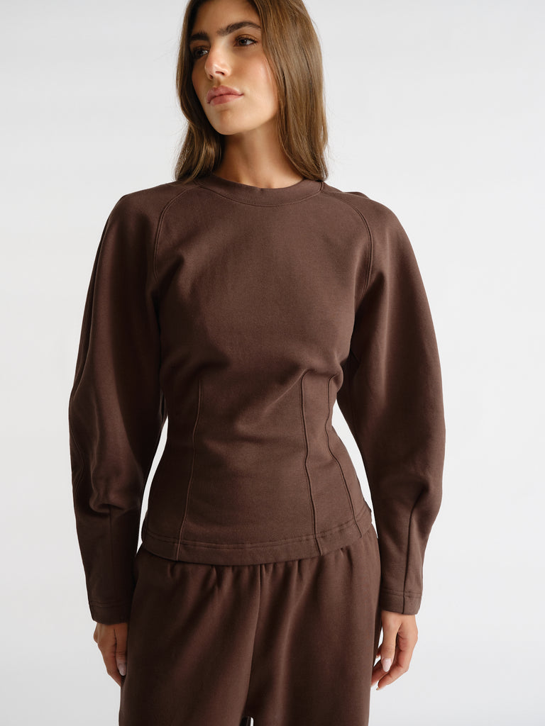 Woman wearing a rich brown cinched waist sweatshirt with long sleeves, paired with matching relaxed pants on a white background