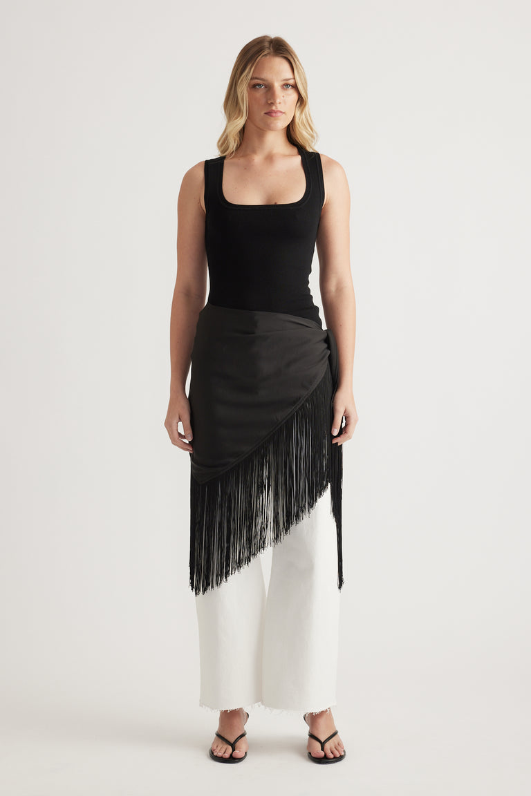 Woman wearing a black square neck knit tank with a fringed skirt, white pants and sandals