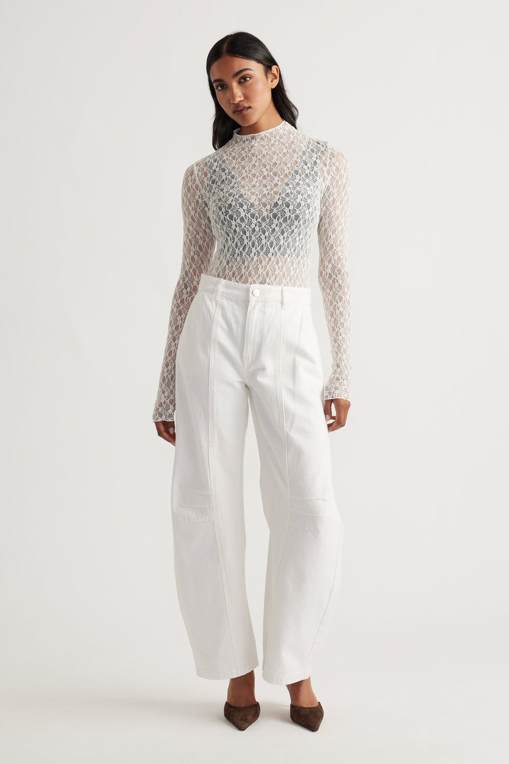 Woman wearing a sheer lace long sleeve top layered over a black bralette paired with high-waisted wide-leg white pants and brown heels