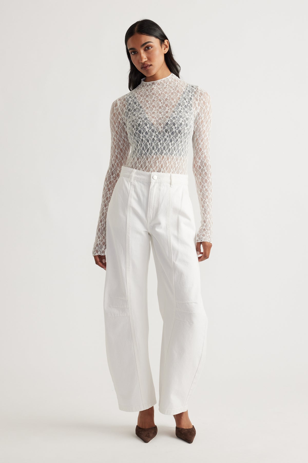 Woman wearing a sheer lace long sleeve top layered over a black bralette paired with high-waisted wide-leg white pants and brown heels