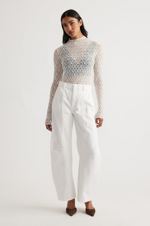 Woman wearing a sheer lace long sleeve top layered over a black bralette paired with high-waisted wide-leg white pants and brown heels