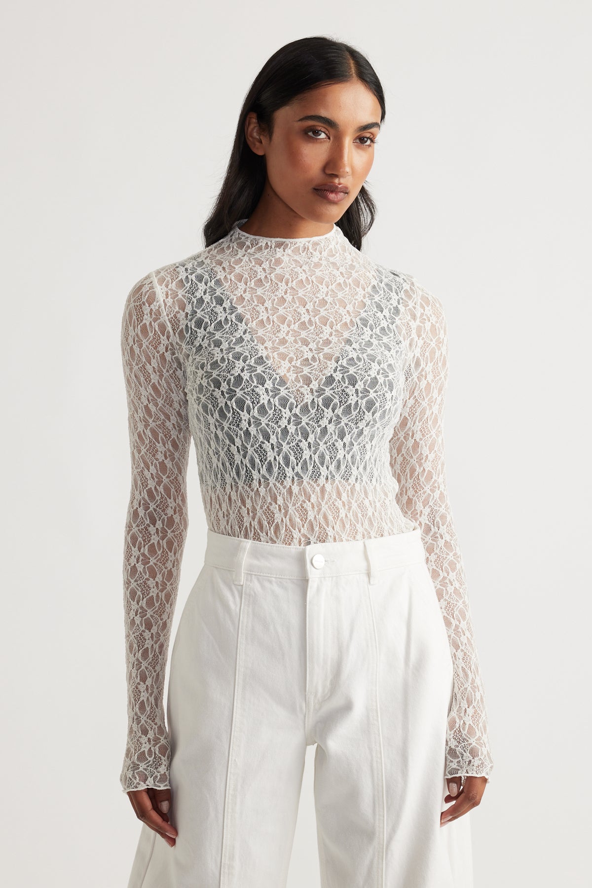 Woman wearing a delicate long sleeve lace top layered over a dark bralette paired with high-waisted ivory pants