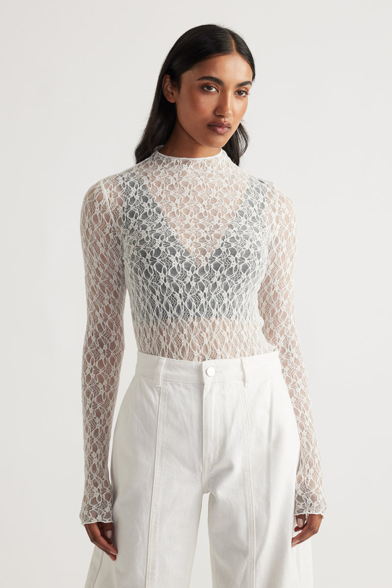 Woman wearing a delicate long sleeve lace top layered over a dark bralette paired with high-waisted ivory pants