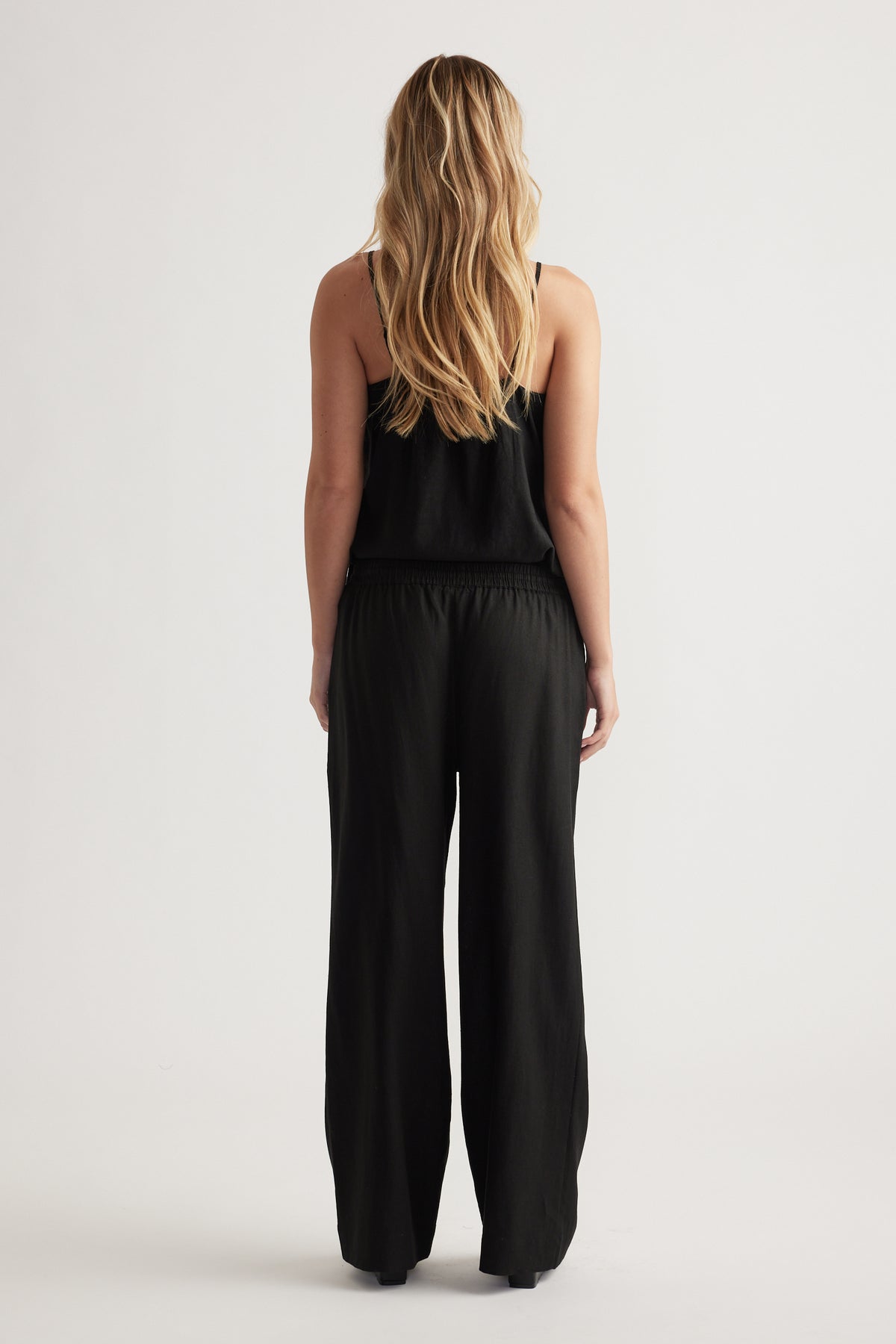 Woman with long blonde hair wearing loose black linen blend pants and a sleeveless black top, standing facing away