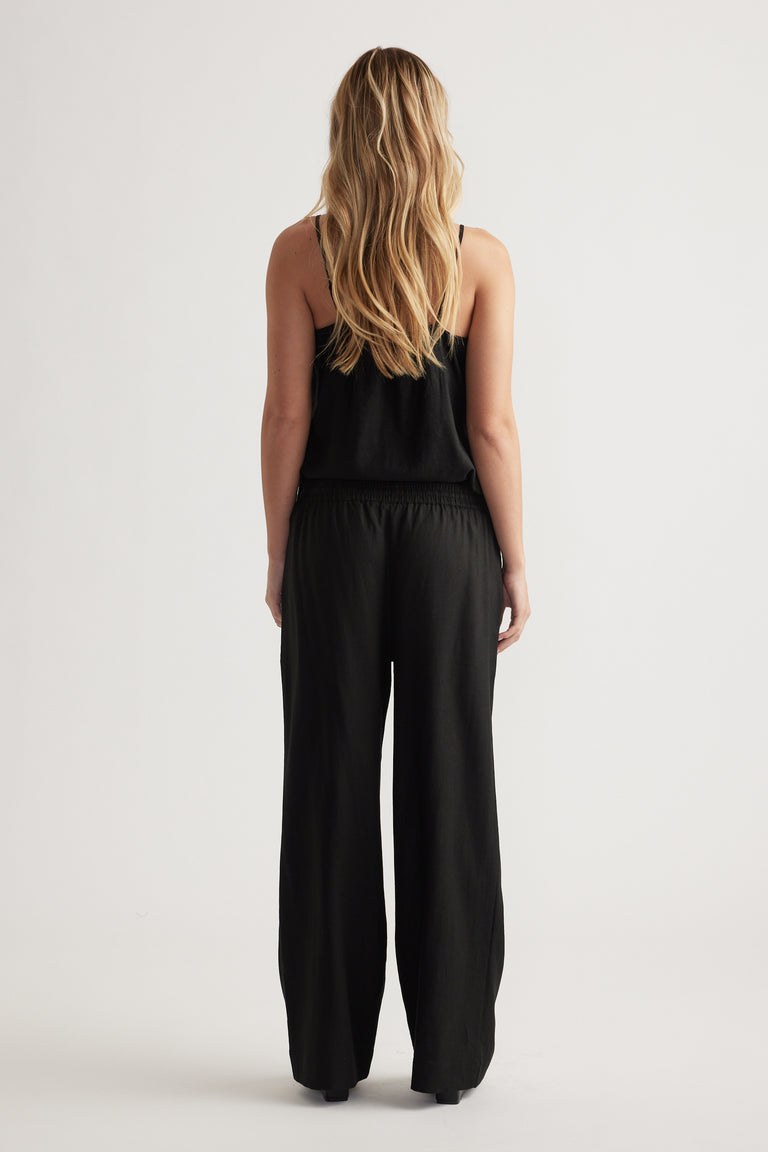 Woman with long blonde hair wearing loose black linen blend pants and a sleeveless black top, standing facing away