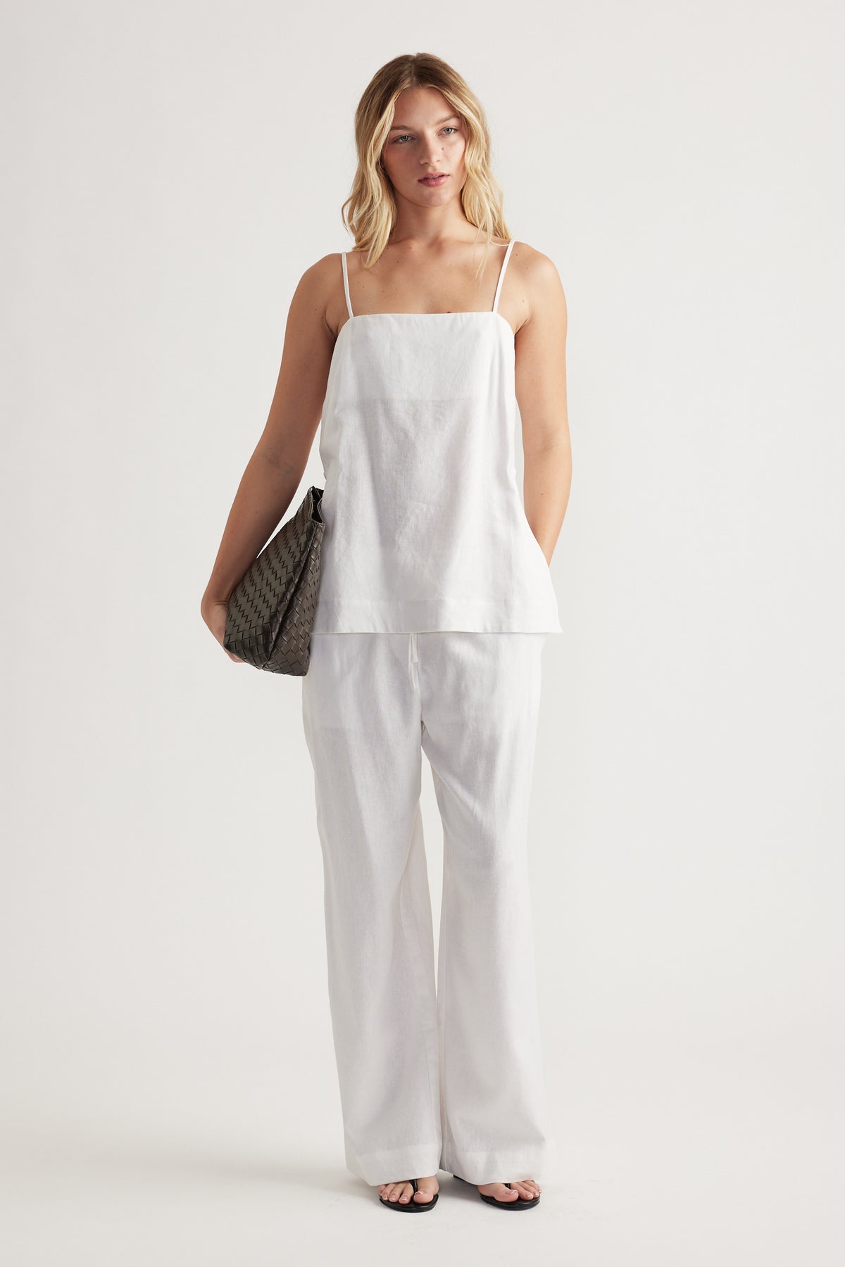Woman wearing relaxed white linen pants paired with a sleeveless square-neck cami and black sandals, holding a patterned bag.