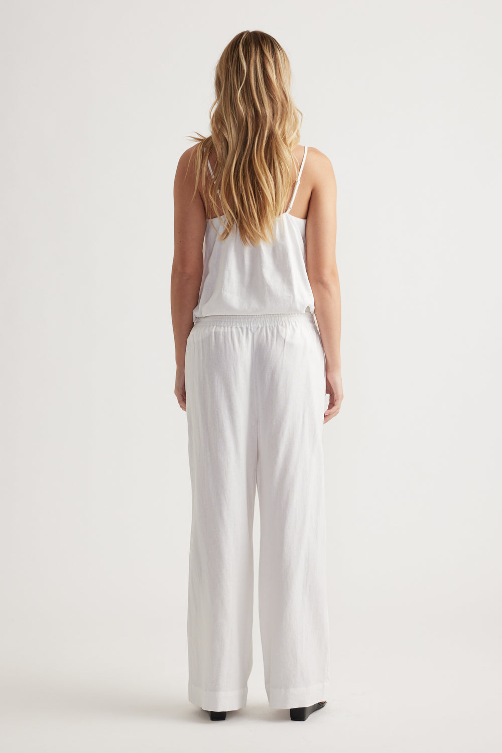 Woman wearing relaxed fit white linen blend pants with elastic waistband and a white sleeveless top, shown from behind