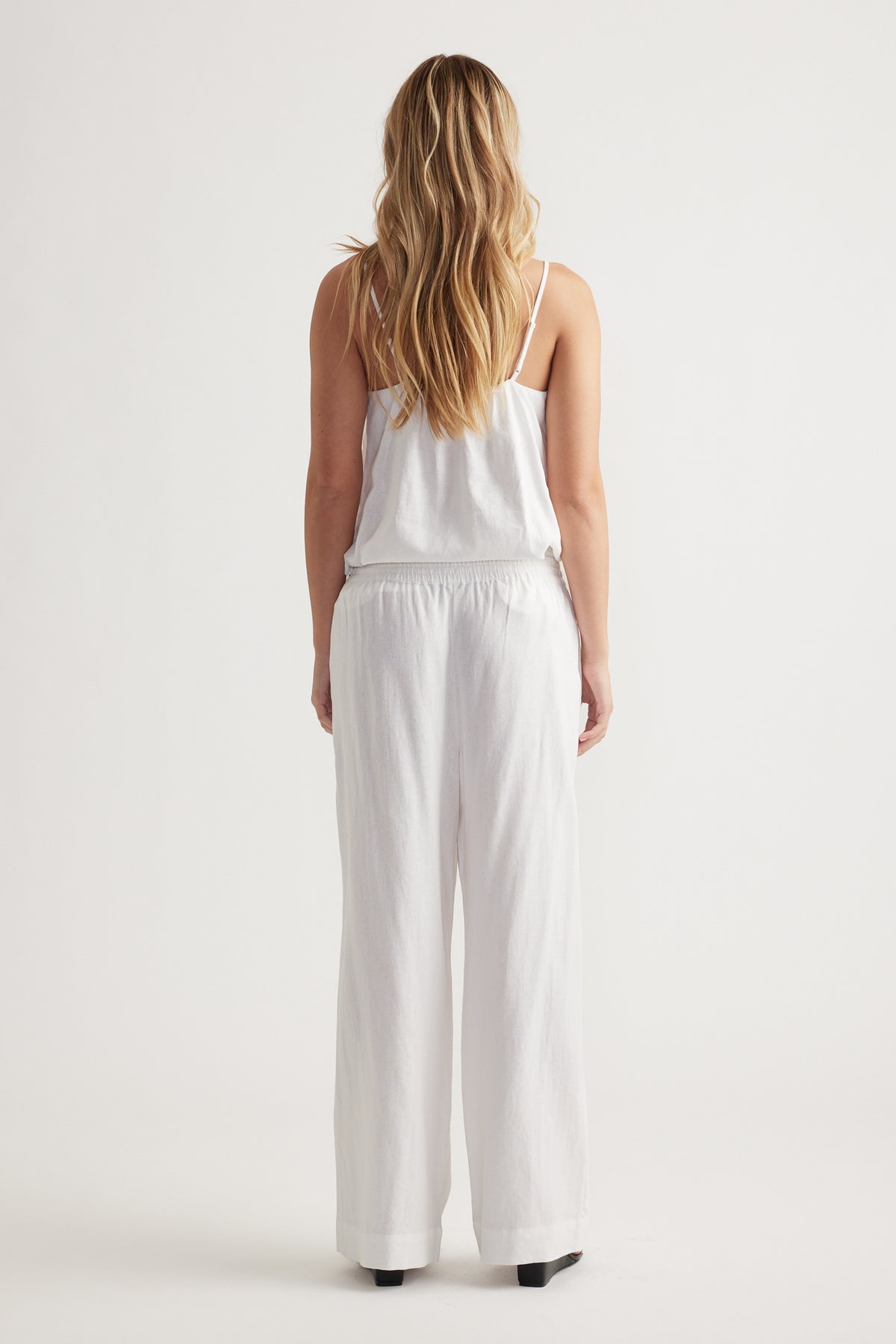 Woman wearing relaxed fit white linen blend pants with elastic waistband and a white sleeveless top, shown from behind