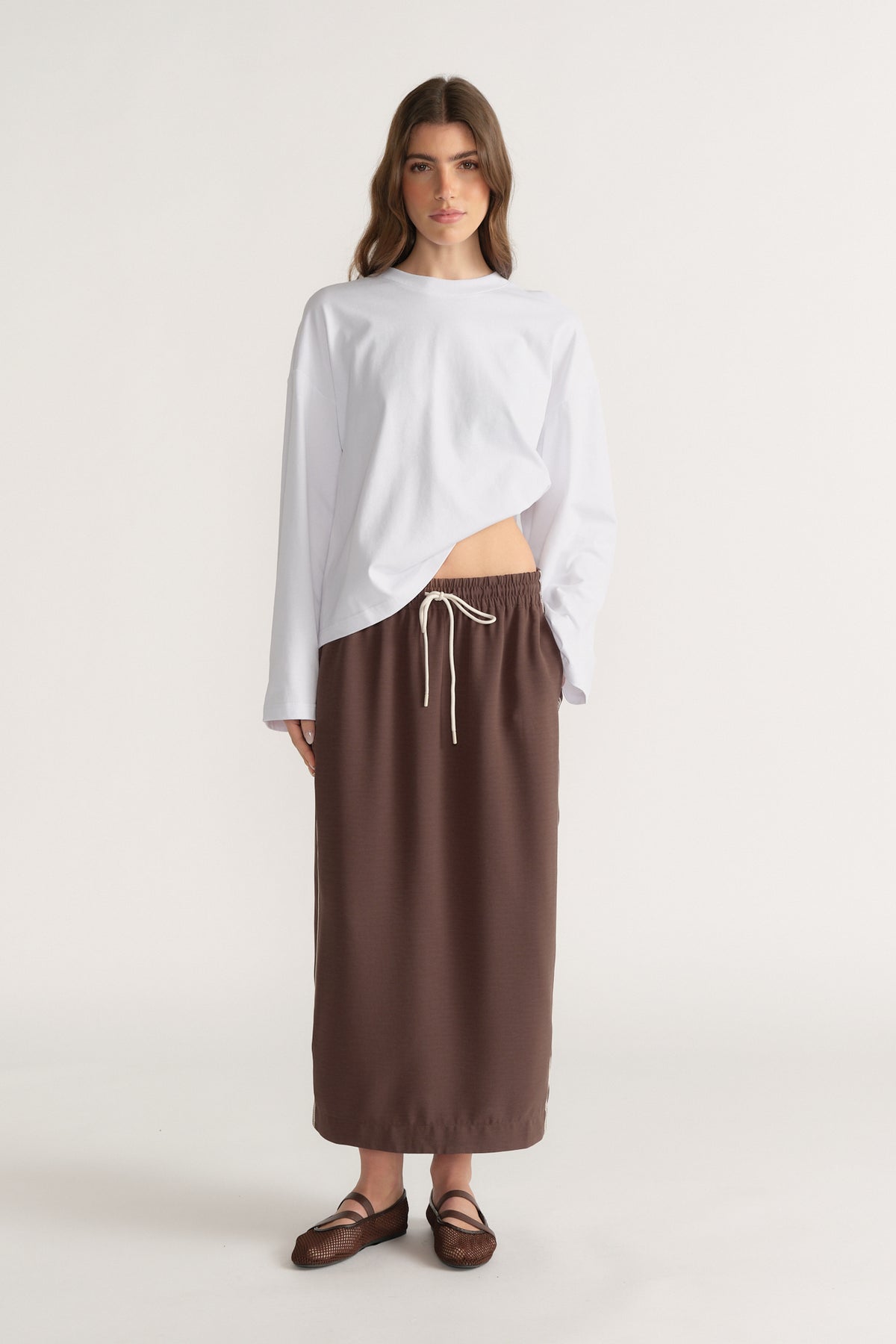 Woman in a brown midi skirt with a drawcord waist, white long sleeve top and strappy sandals