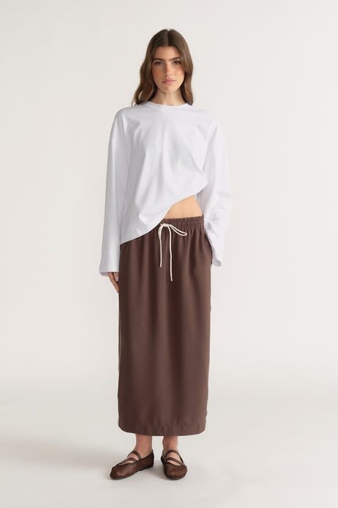 Woman in a brown midi skirt with a drawcord waist, white long sleeve top and strappy sandals
