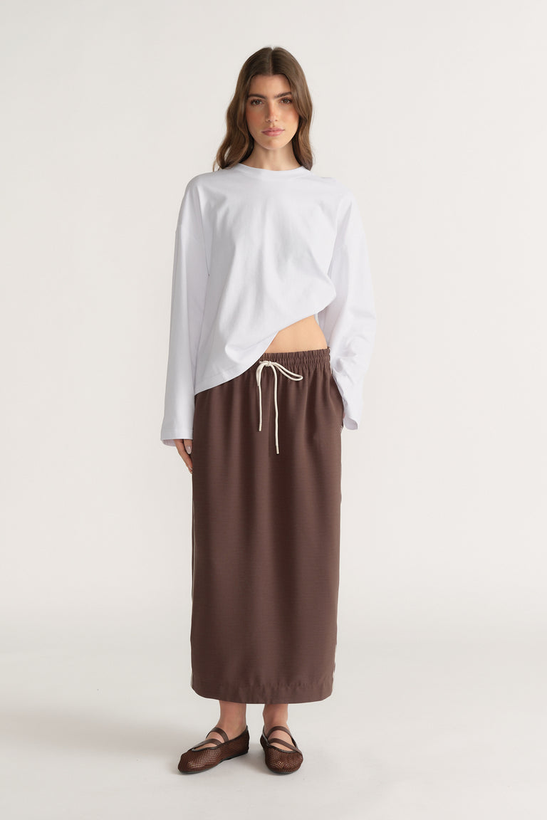 Woman in a brown midi skirt with a drawcord waist, white long sleeve top and strappy sandals
