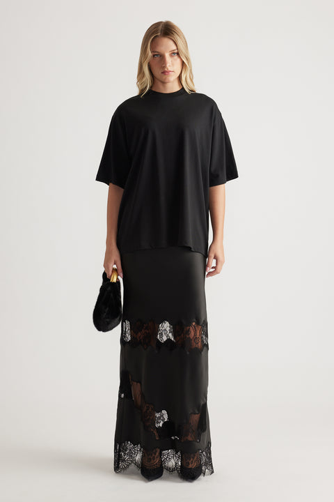 Woman wearing a loose black oversized tee paired with a long black skirt featuring lace panels and holding a small black bag