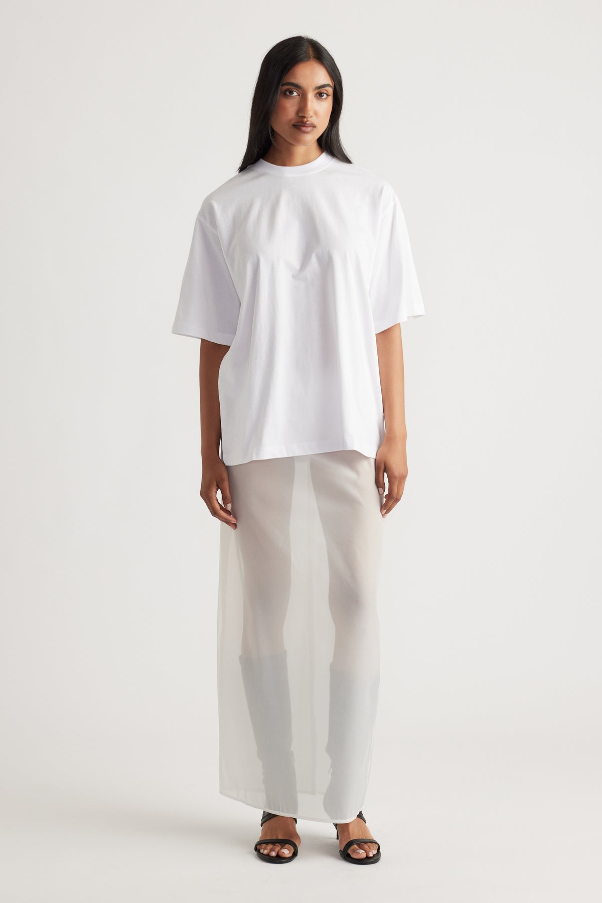 Woman wearing a relaxed white oversized tee paired with a sheer maxi skirt and black open-toe sandals for a modern look