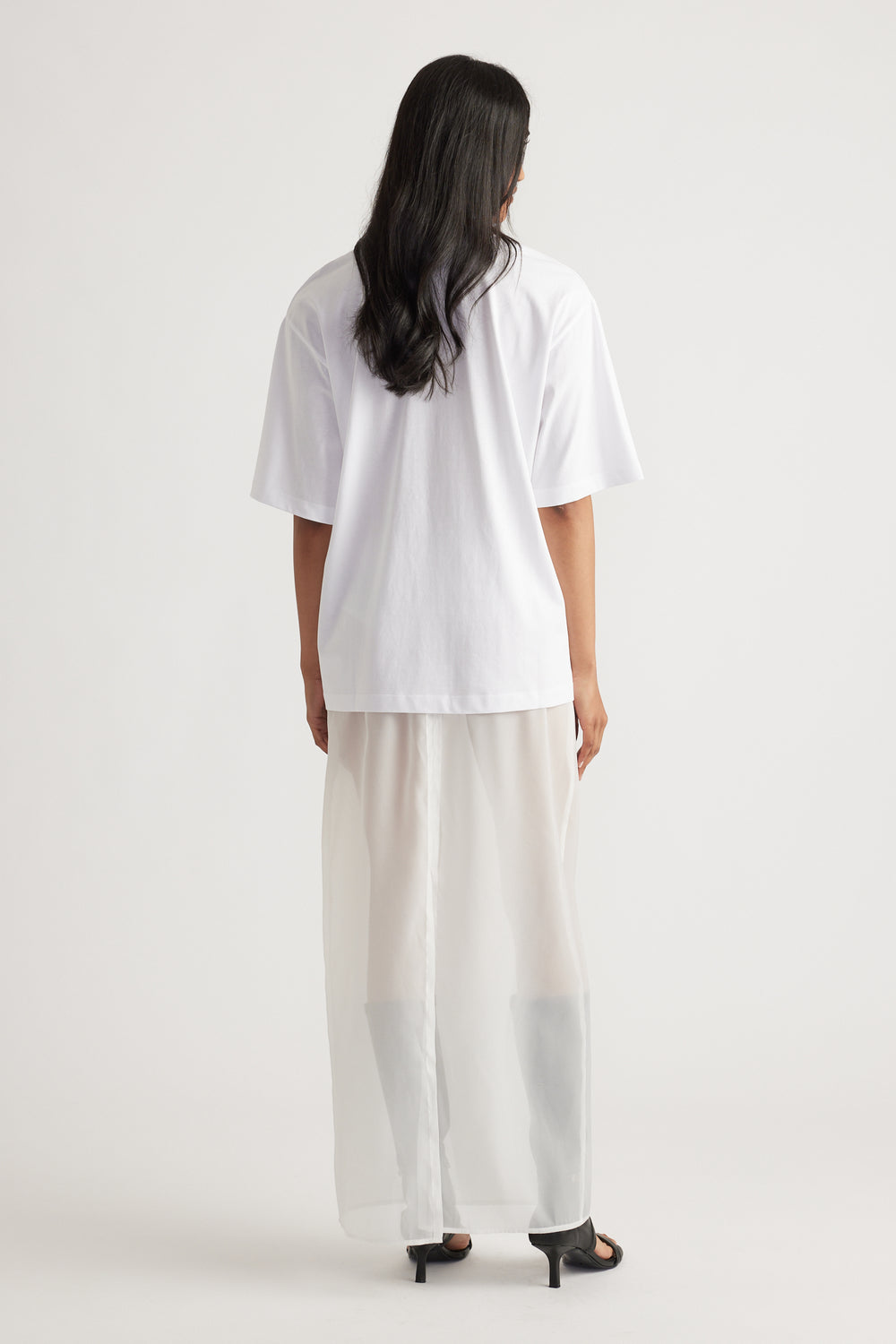 Rear view of woman wearing a loose white oversized tee paired with wide-leg sheer cream pants and black heels