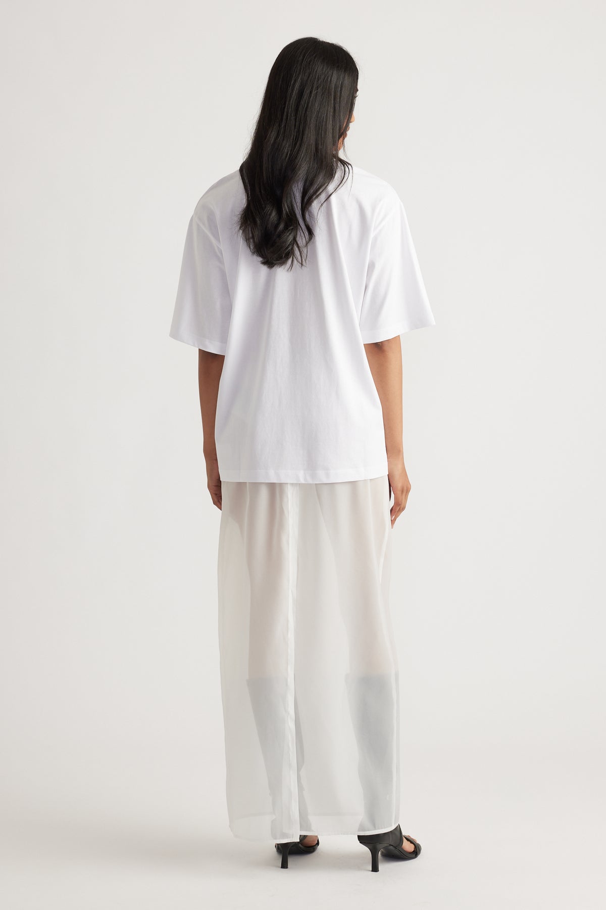 Rear view of woman wearing a loose white oversized tee paired with wide-leg sheer cream pants and black heels