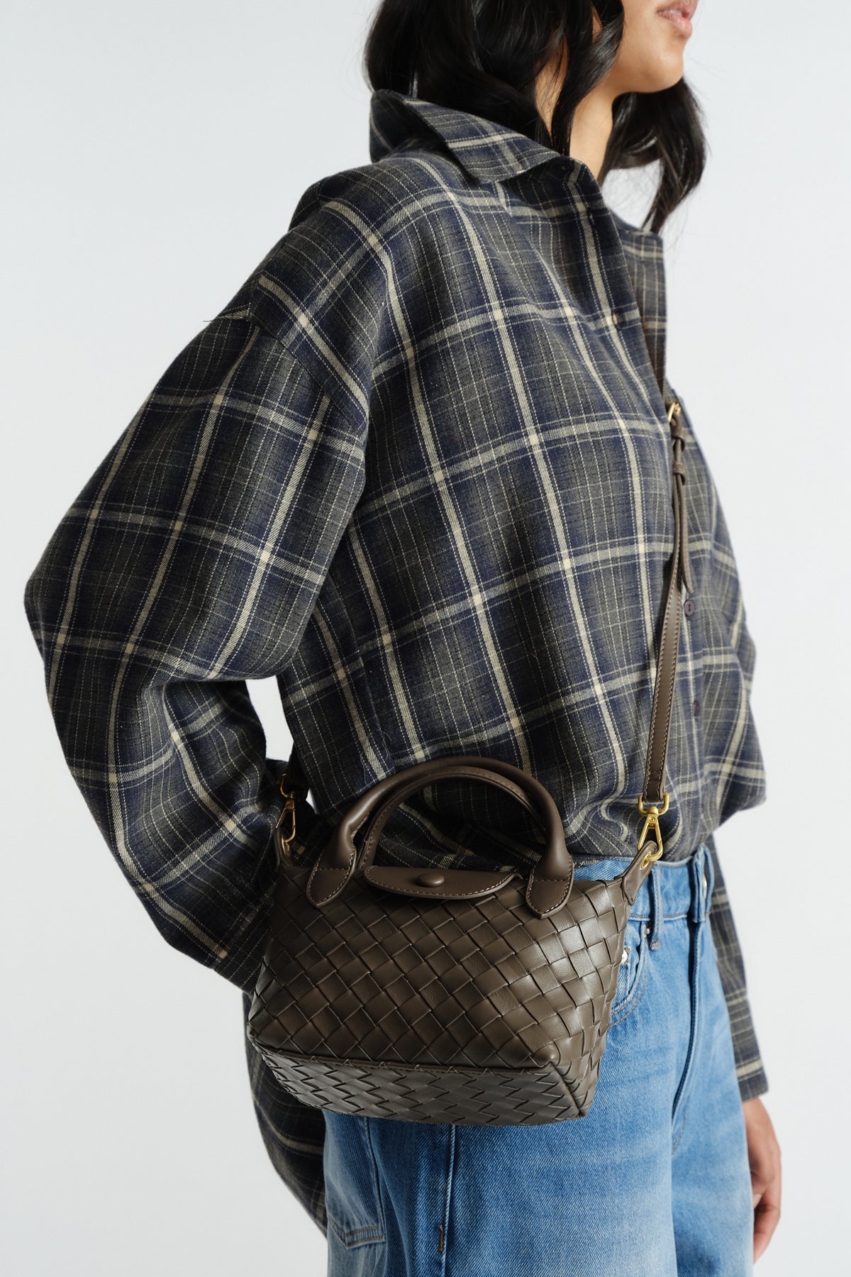 Woven leather mini bag in dark khaki with top handle and crossbody strap, styled with casual plaid shirt and denim jeans.