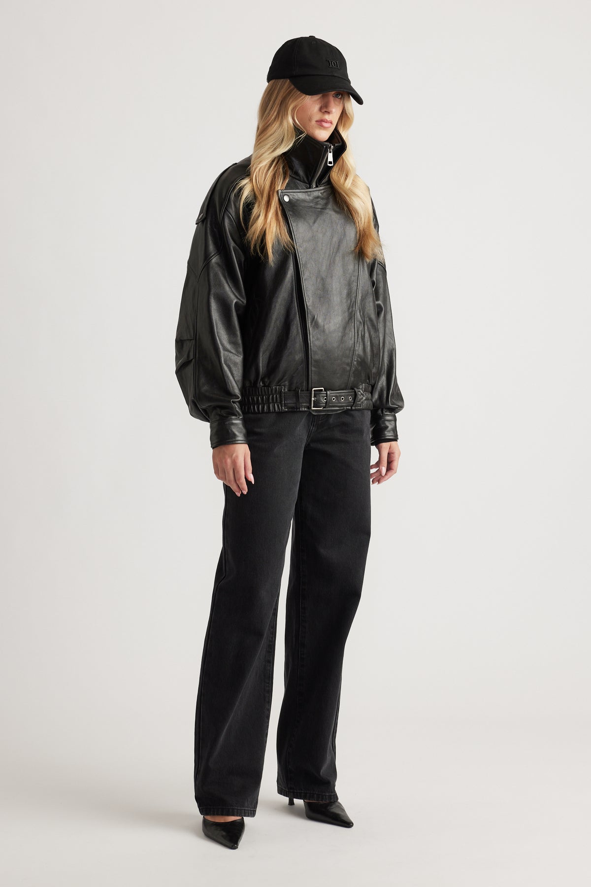 Woman wearing oversized black leather jacket with belt detail, wide-leg black jeans, pointed heels, and a black vintage cap