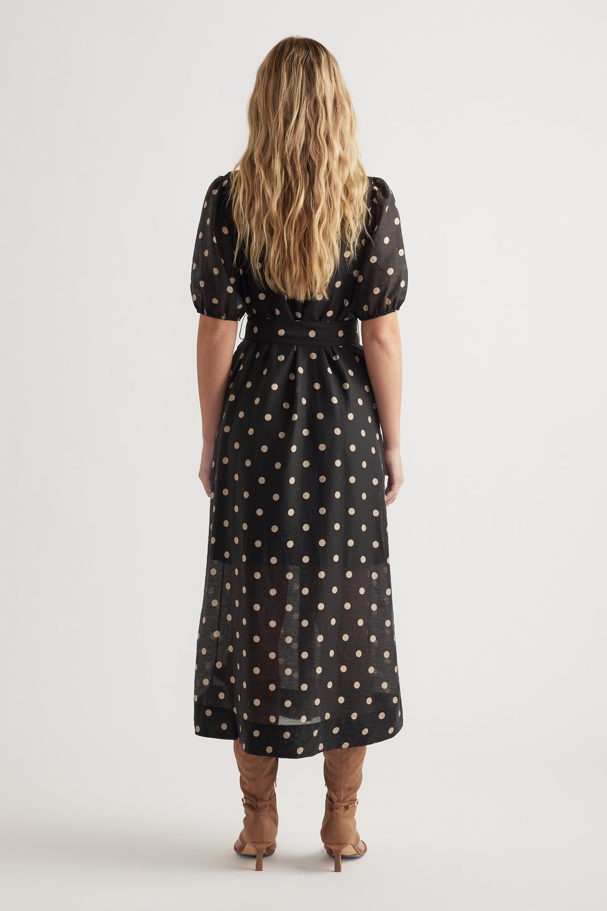 Back view of woman wearing a black midi dress with puff sleeves and beige polka dots paired with tan boots.