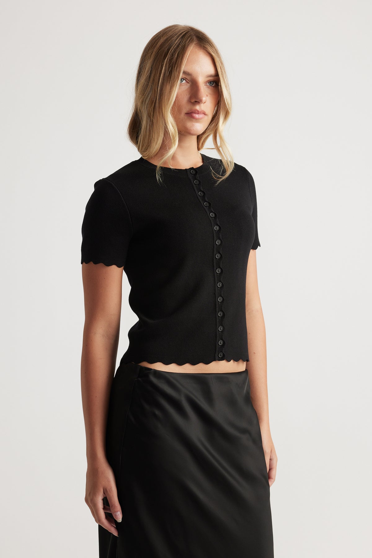 Woman wearing black Maya Scallop Trim Button Top with short sleeves and scalloped edges, paired with a sleek black skirt