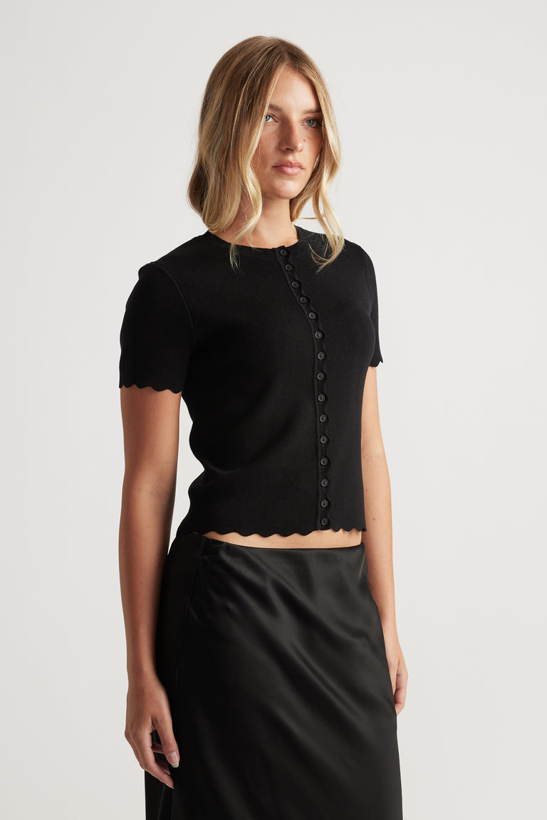Woman wearing black Maya Scallop Trim Button Top with short sleeves and scalloped edges, paired with a sleek black skirt