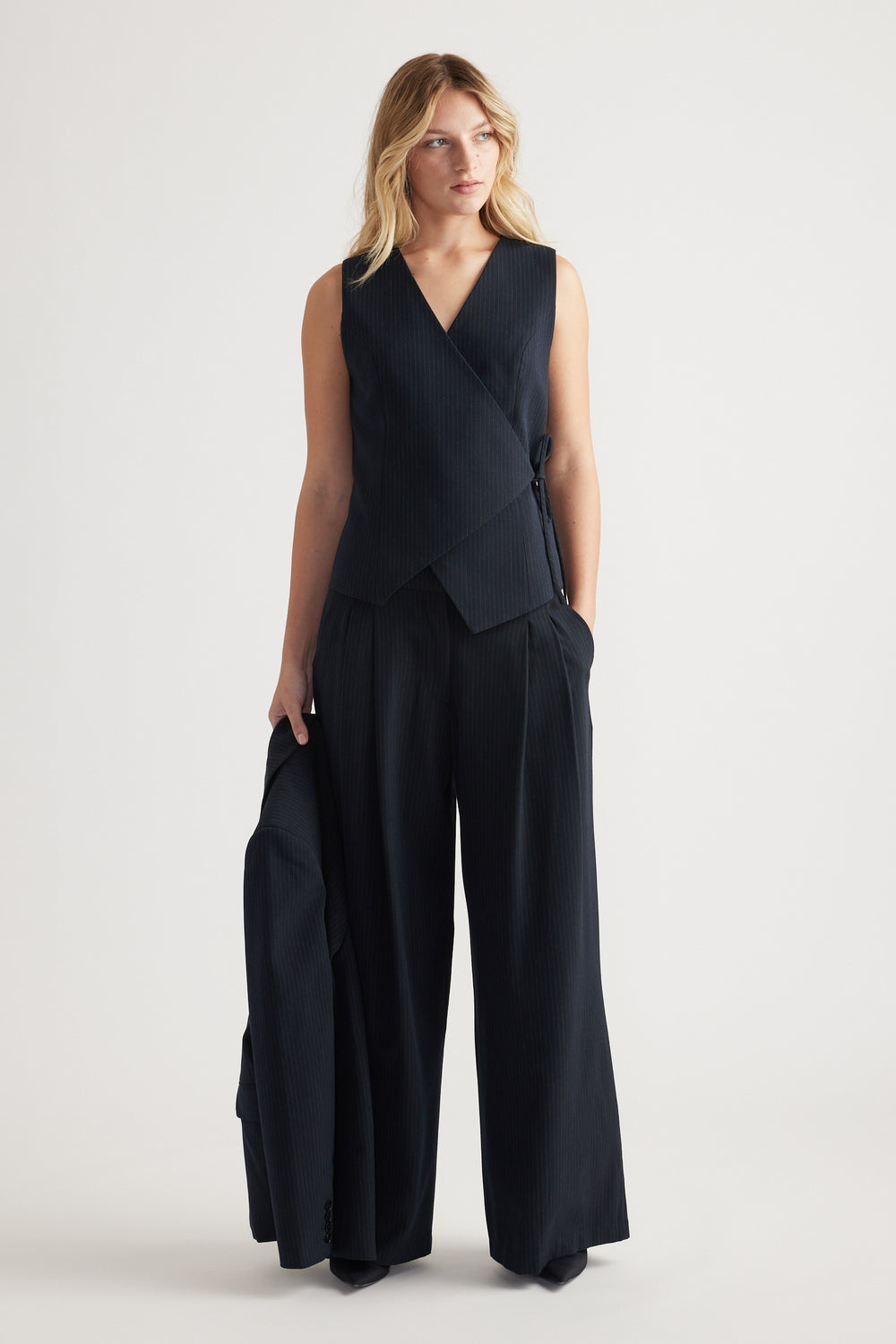 Woman wearing a navy wrap vest with side tie detail paired with wide-leg trousers, holding a matching blazer.