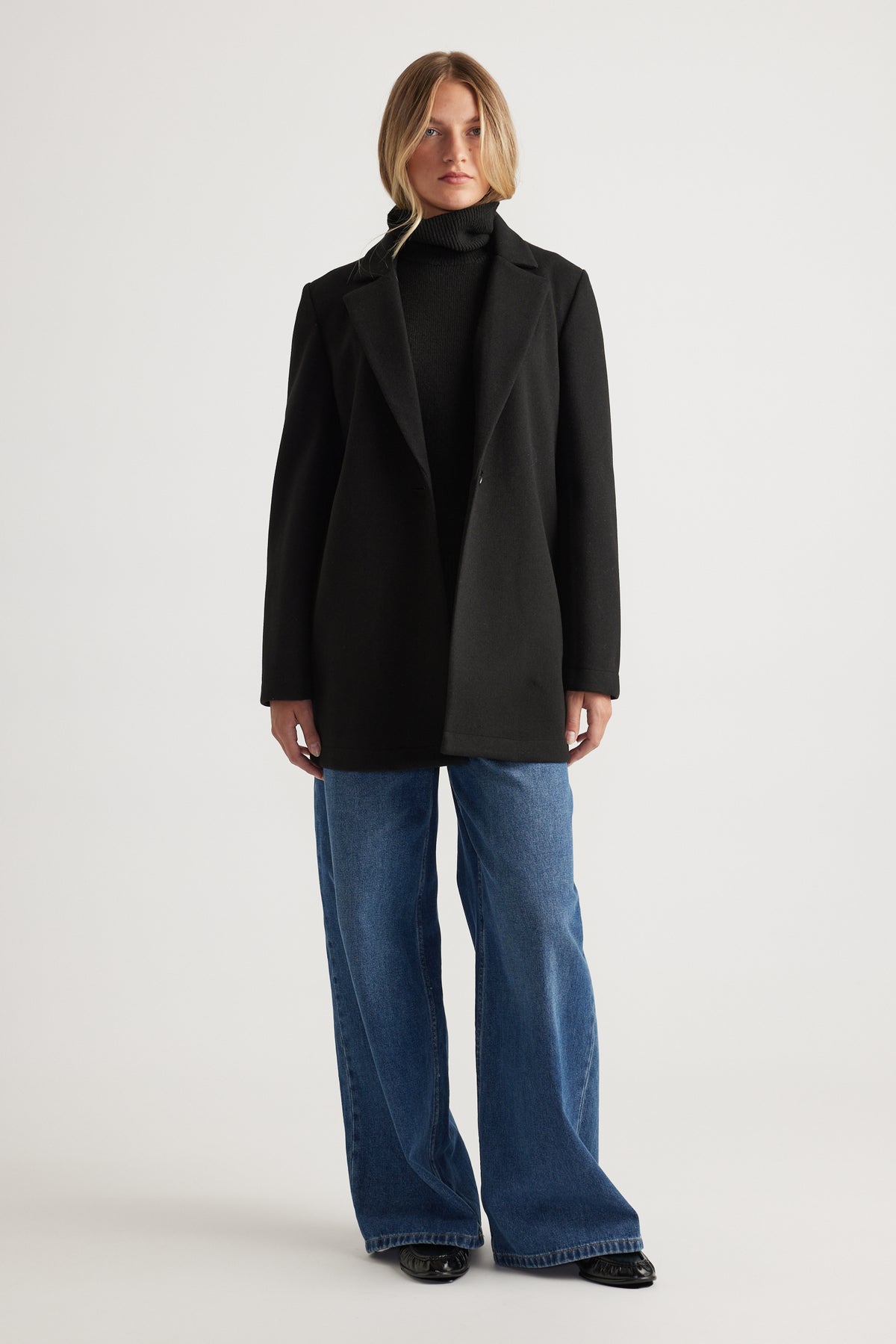 Woman wearing a classic black mid length felt coat over a black turtleneck paired with wide-leg blue jeans and black shoes