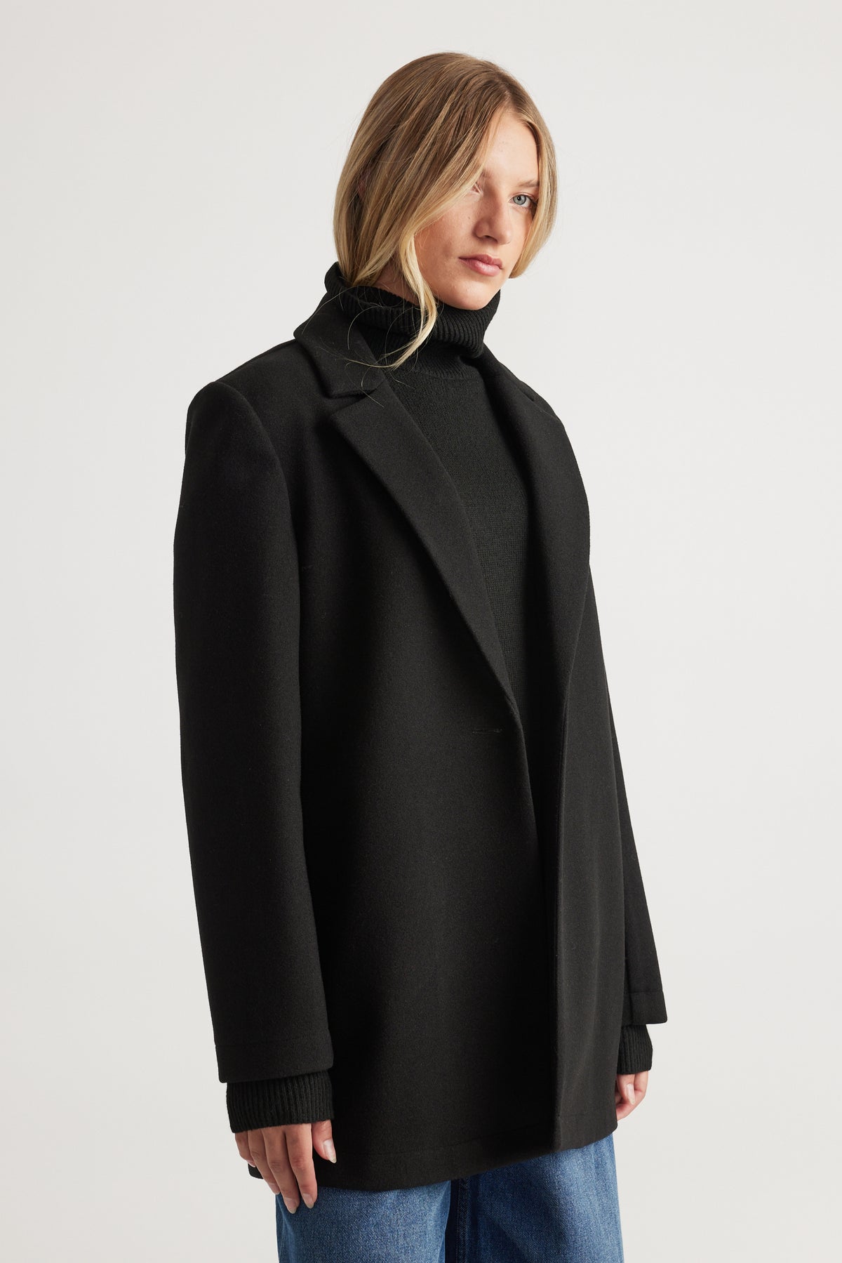Woman wearing a sleek black mid-length felt coat layered over a black turtleneck and blue denim jeans.